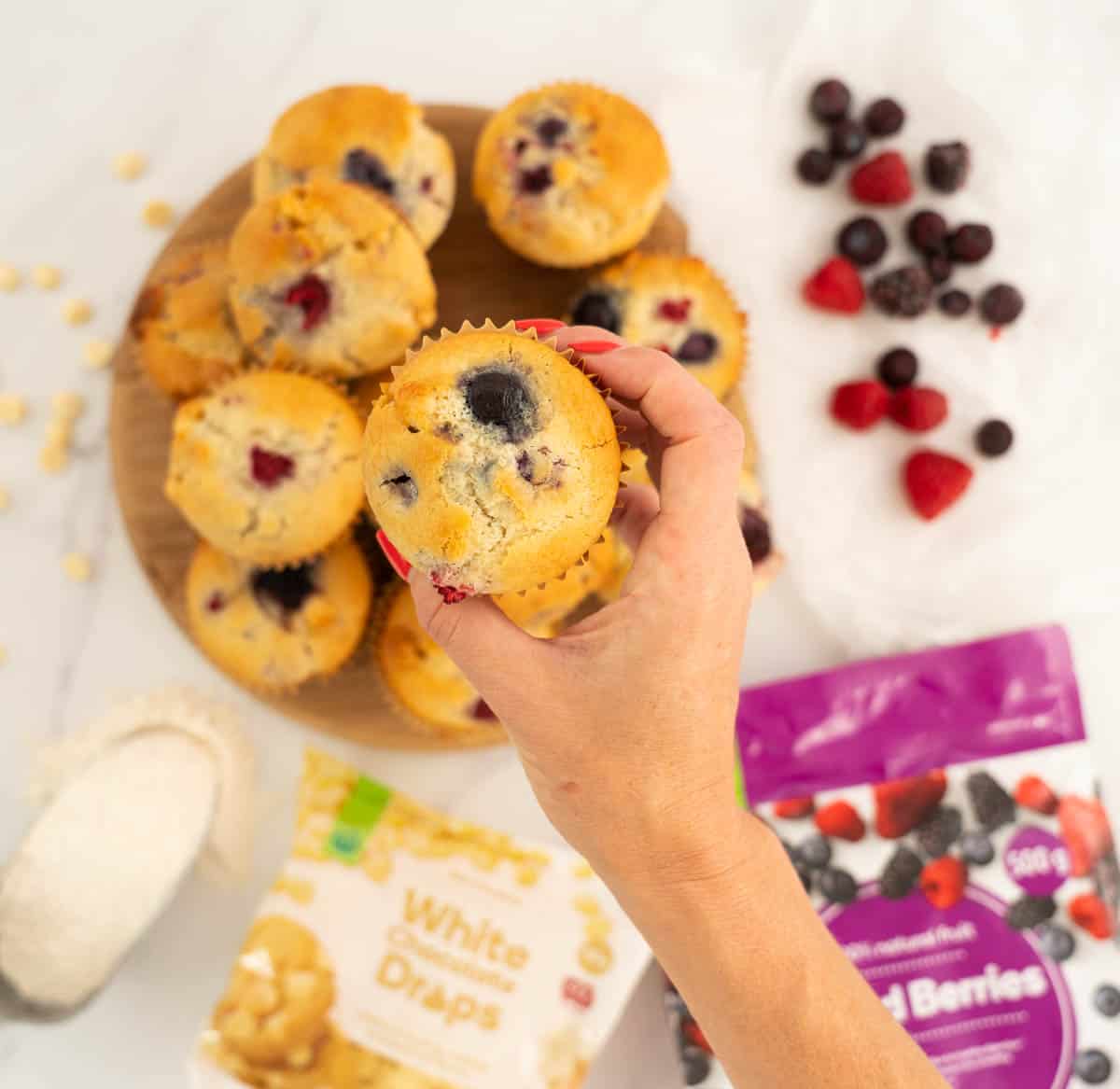 Woman's hand holding a berry muffin above a wooden tray of muffins.