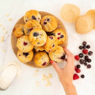 Women's hand reaching for a berry muffin from a wooden tray of a dozen muffins.