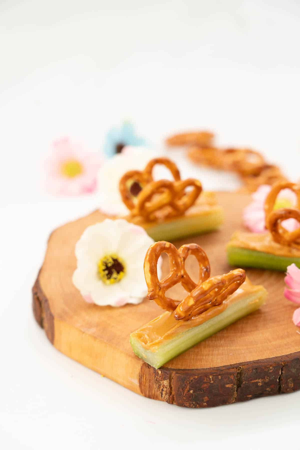 Celery sticks filled with peanut butter decorated with pretzels to form wings, making a butterfly snack., on a wooden board scattered with small flowers.