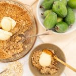 Feijoa crumble in glass dish with serving spoon inside and dessert bowl with serving of feijoa crumble and spoon inside. Bowl of fresh feijoas in bowl on benchtop.