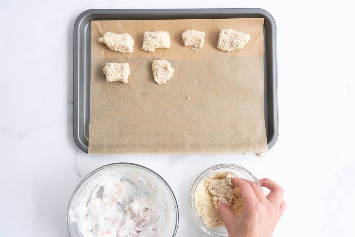 Baking paper lined tray with chicken nuggets on it, next to a women's hand rolling chicken pieces in ground almond.