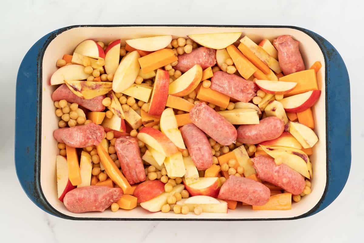 Sliced apples, carrots, sweet potato, sausages and chickpeas in a large blue ceramic roasting pan.