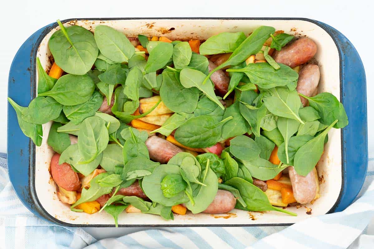 Spinach leaves on top of roasted vegetables and sausages in a blue ceramic roasting pan.