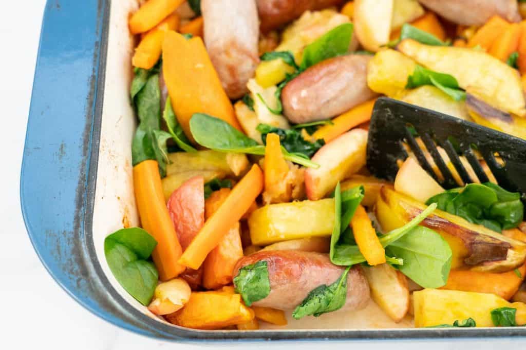 Close up of Baked sausages, apples, sweet potato and spinach leaves in a blue ceramic roasting pan.