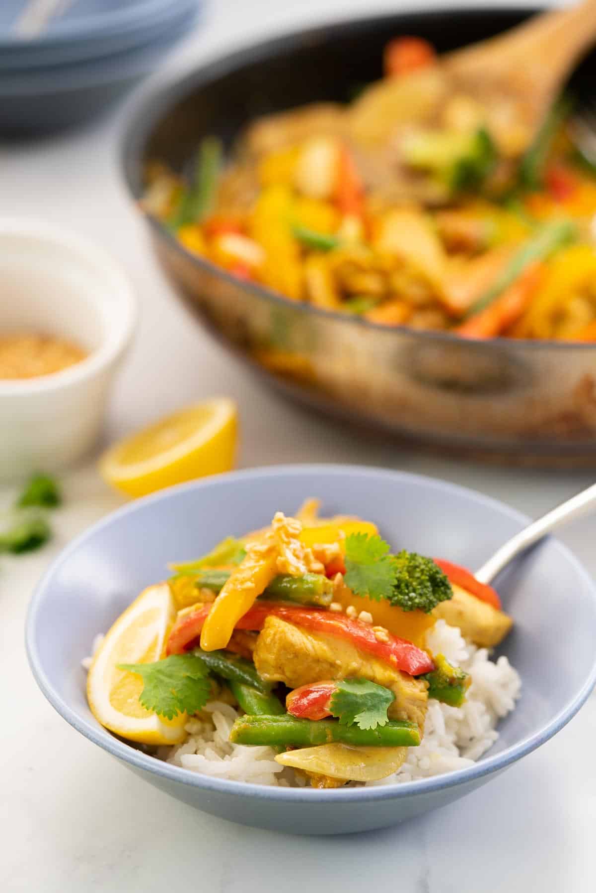 A bowl of chicken satay stir fry on a bed of rice, with a large fry pan in the background.