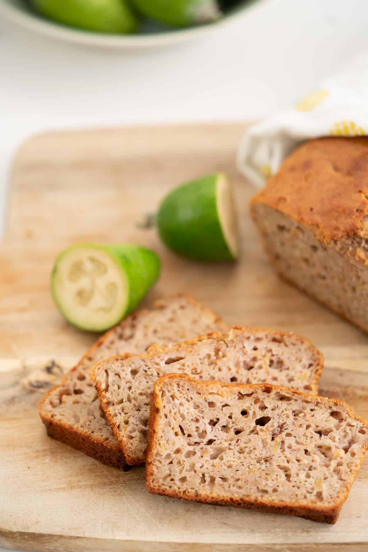 Three slices of loaf on a wooden chopping board with sliced feijoas in the background.