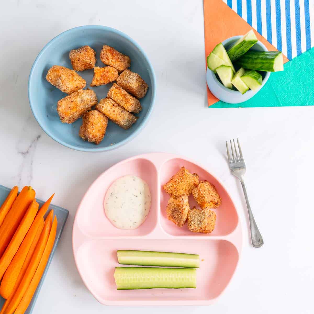 Baby meal served in a pink divided plate with salmon nuggets, cucumber and dipping sauce.