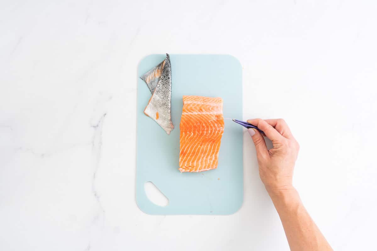A salmon fillet with skin removed, hand using tweezers to remove bones.