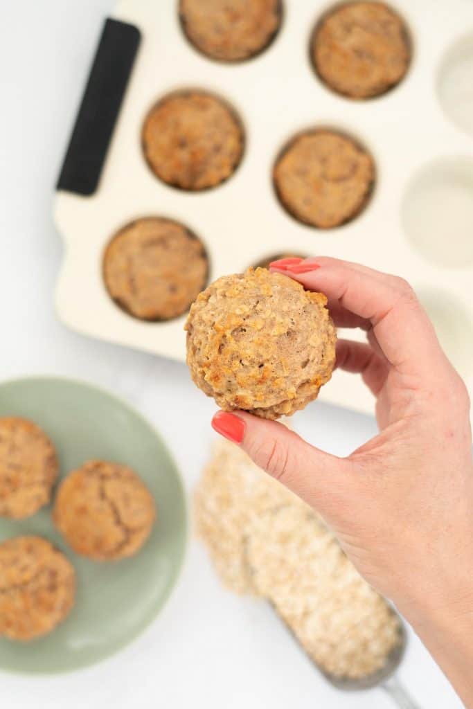 Women's hand holding an apple toddler muffin above a kitchen counter top.