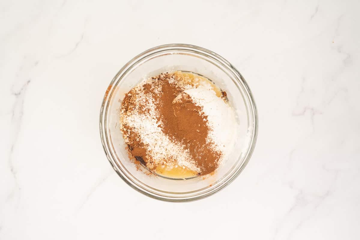 Dry ingredients sprinkled on top of wet ingredients in a medium sized glass mixing bowl.