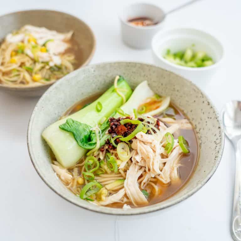 A grey ceramic bowl of chicken noodle soup. chili oil, noodles and bok choy visible.