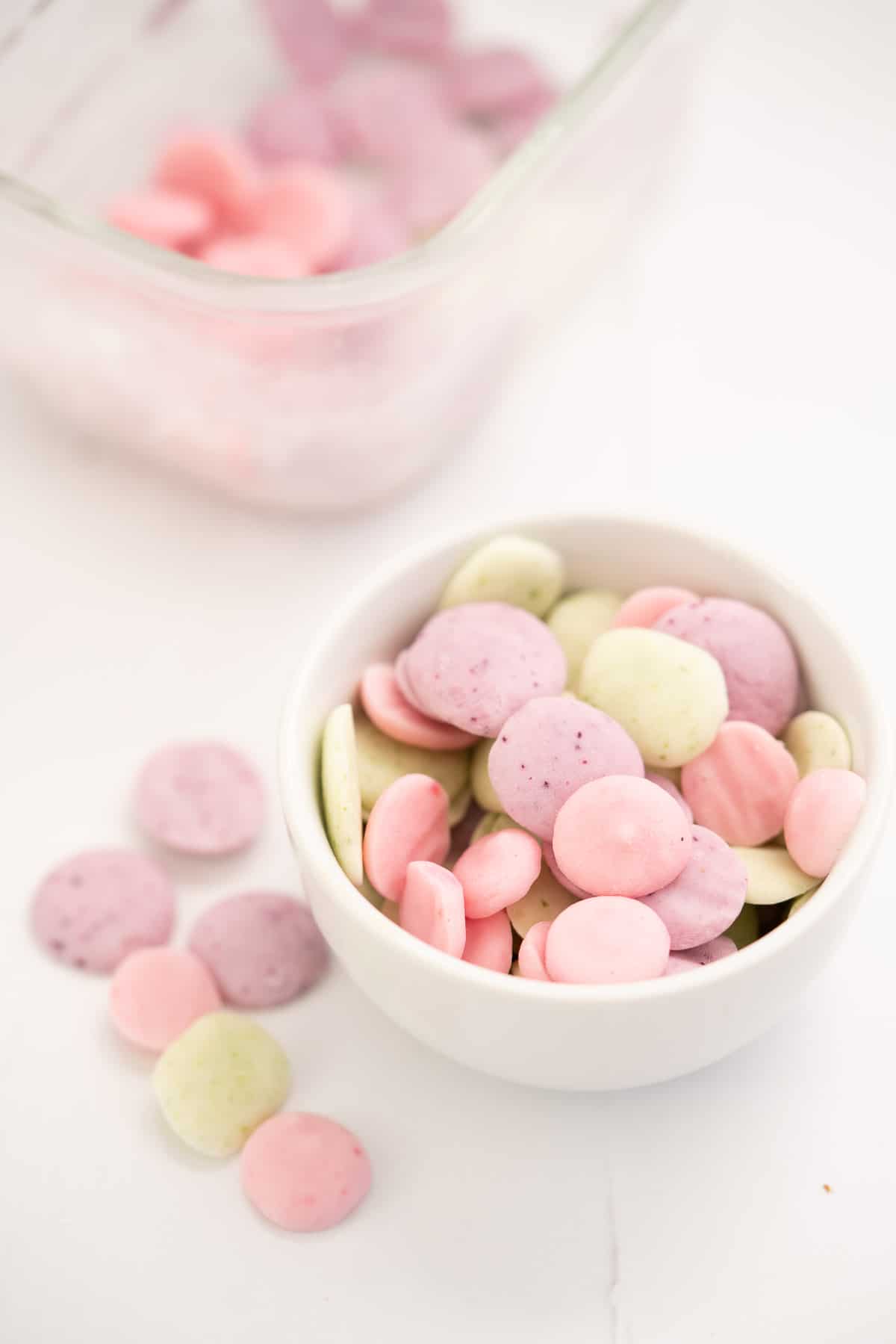 A small white ceramic bowl filled with pink, purple and green baby yogurt melts, a glass container of melts visible in the background.