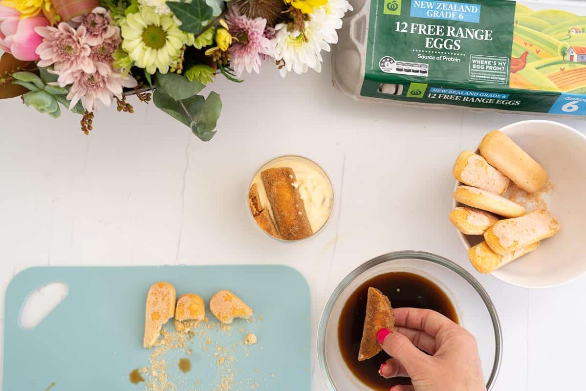 A women's hand holding a piece of coffee dipped savoiardi biscuit ready to layer into a glass.