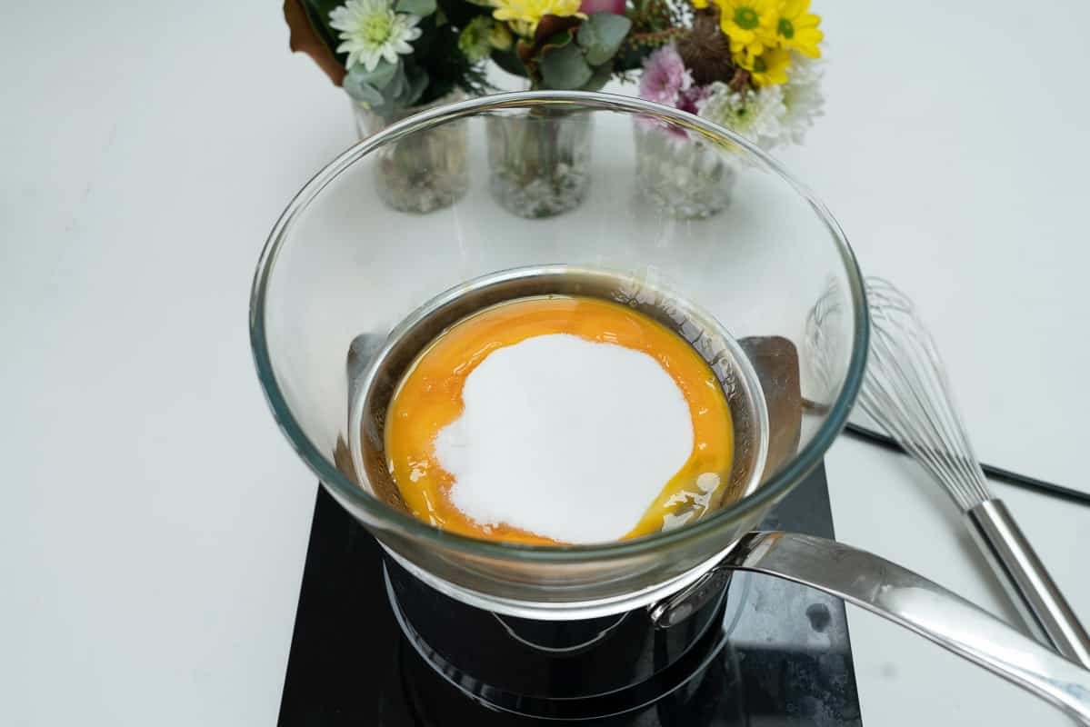 Egg yolks and sugar in a glass bowl sitting on top of a saucepan.