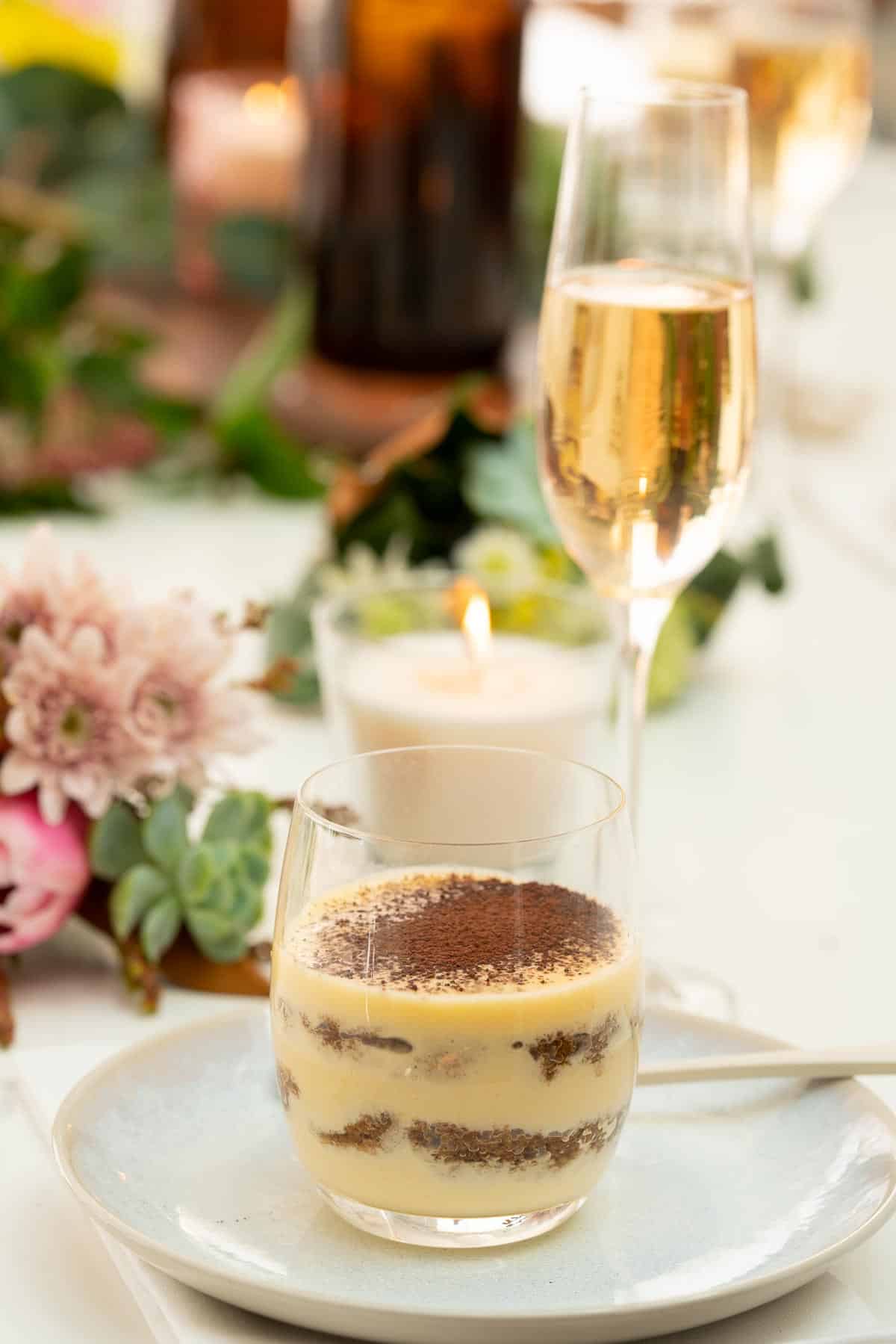 A tiramisu cup sitting on a table decorated with candles and fresh flowers, a glass of champagne and water dcanters visible in the background.