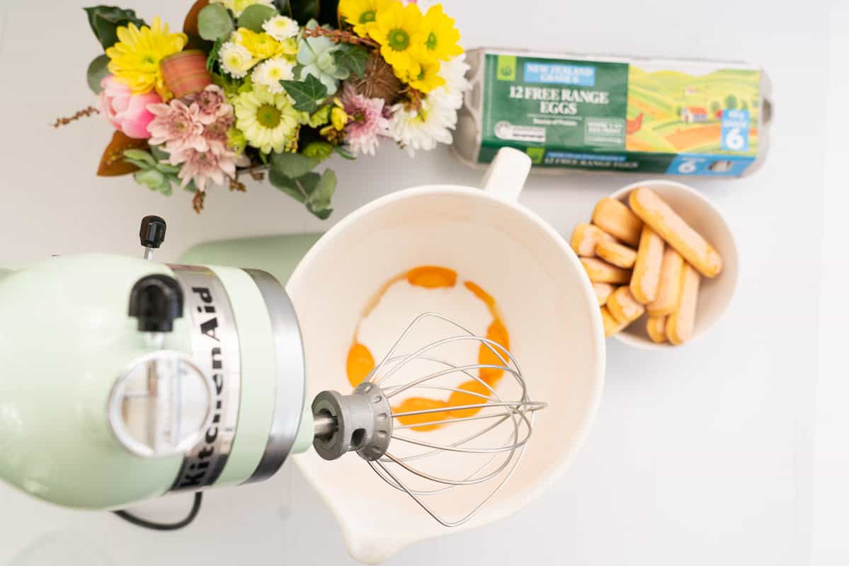 Pale green stand mixer set up with whisk attachment, egg yolks and sugar in the mixing bowl.