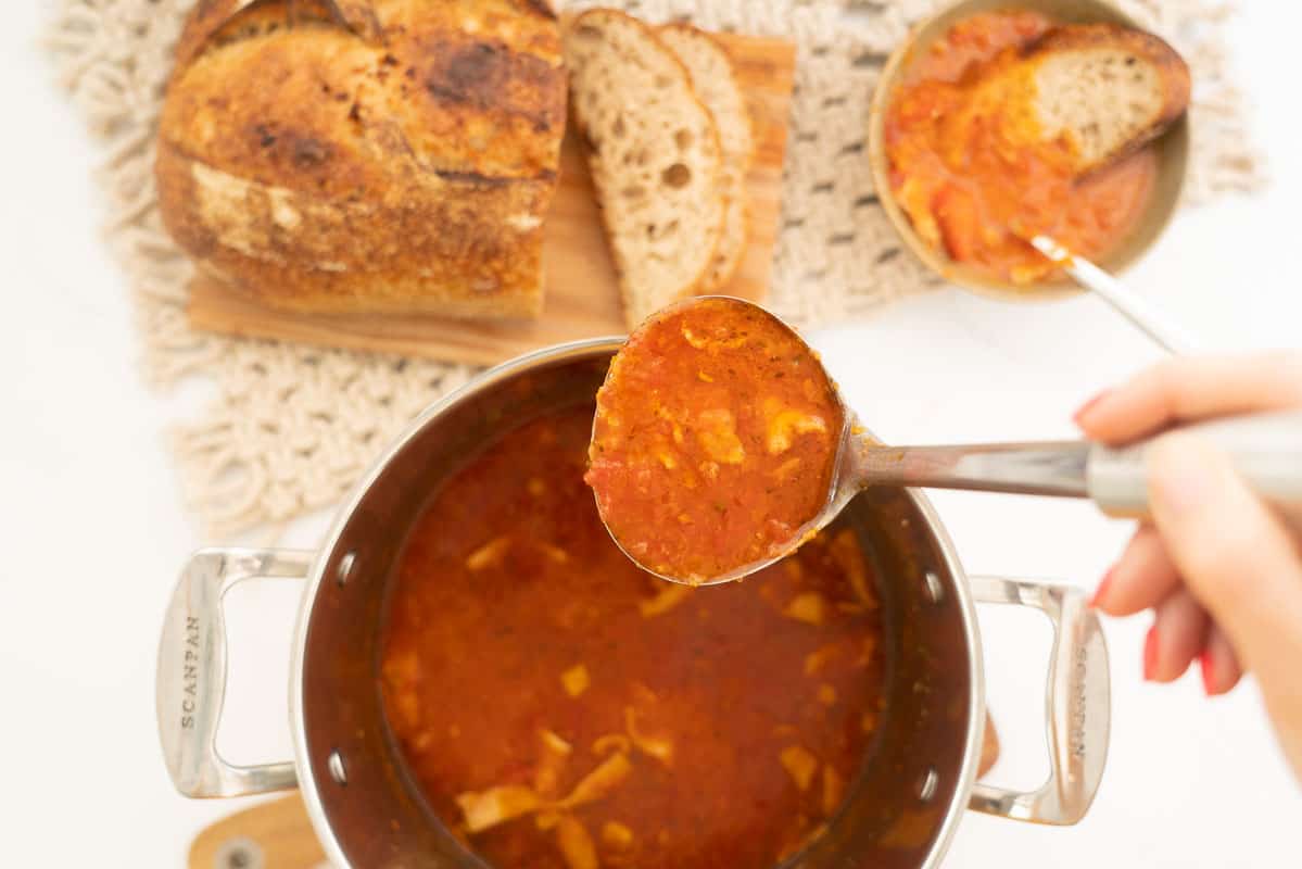 Tomato soup being ladled from a large saucepan into a bowl with crusty bread.