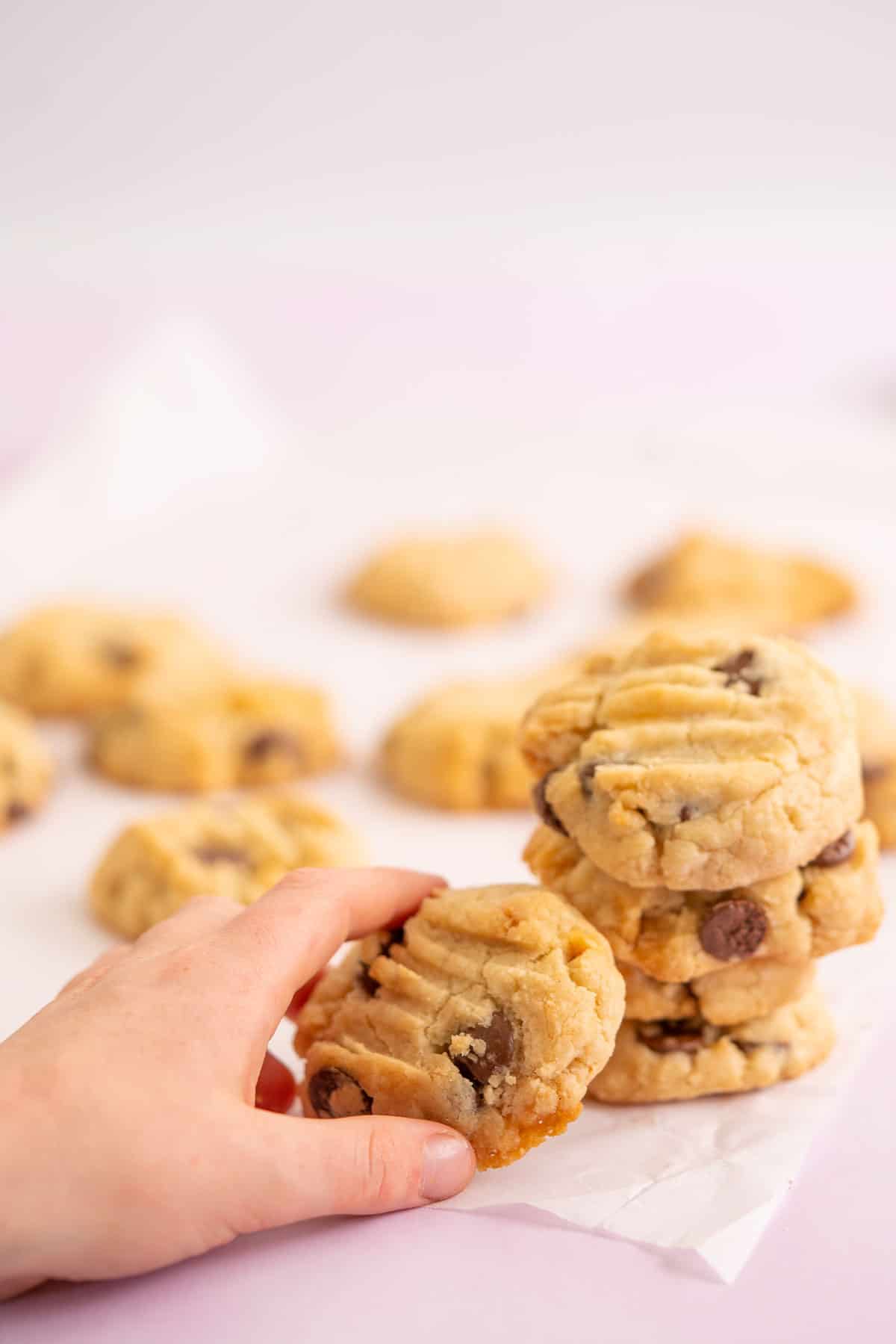 Childs hand reaching for a chocolate chip cookie from a stack of cookies on a pink bench top.