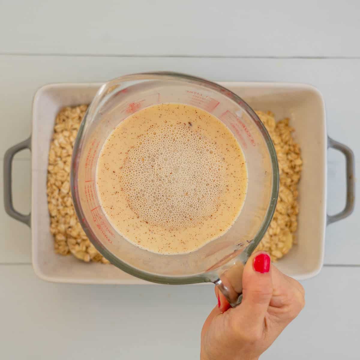 A jug of whisked milk eggs and cinnamon about to be poured over a baking dish of rolled oats.
