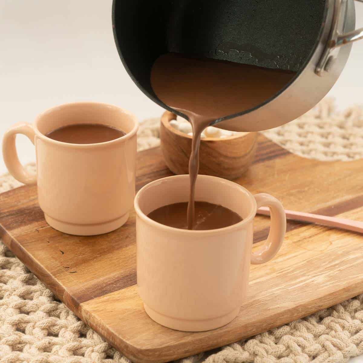 Hot chocolate being poured from a saucepan into a pink mug.