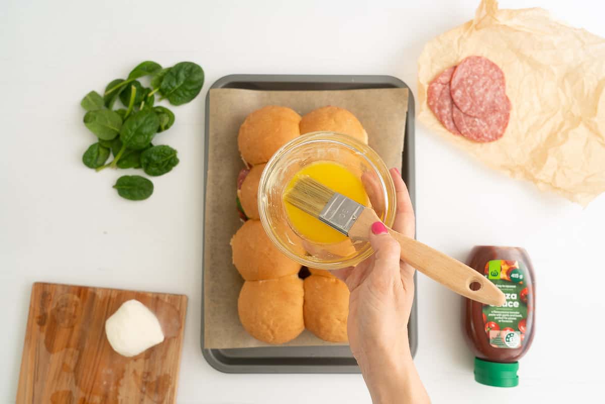 A woman's hand holding a small bowl of melted butter and pastry brush above eight small buns.