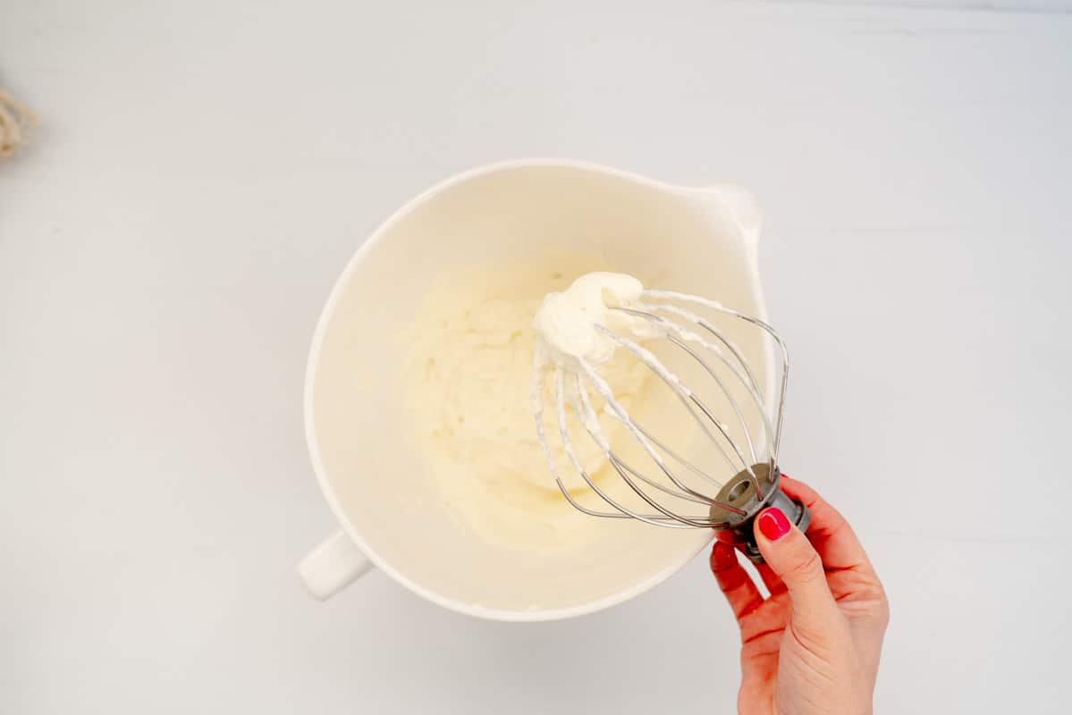 A woman's hand holding the whisk attachment of a stand beater covered in whipped cream.