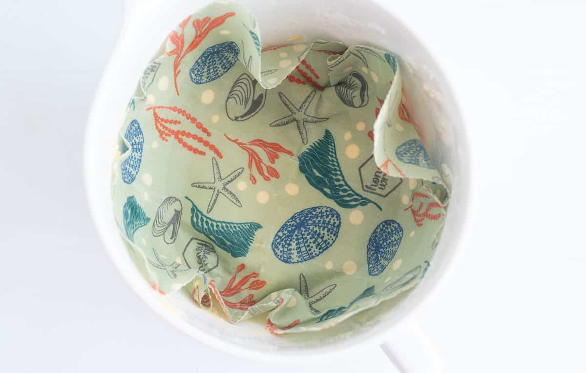 A beeswax wrap pushed down on top of cookie dough in a white ceramic mixing bowl.