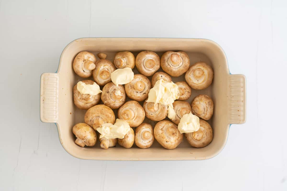 A rectangular baking dish filled with olive oil coated button mushrooms. topped with five blobs of creme fraiche.