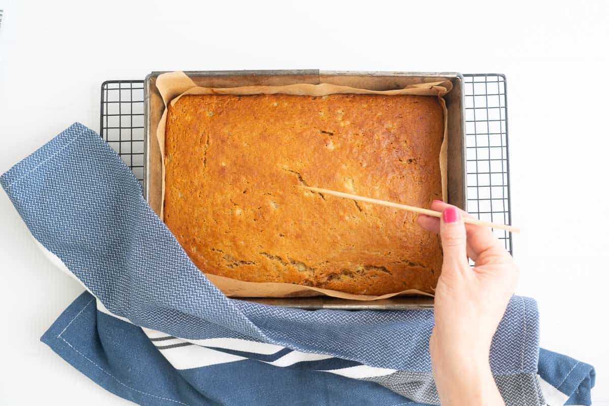 A woman's hand holding a bamboo skewer above a cooked banana cake.
