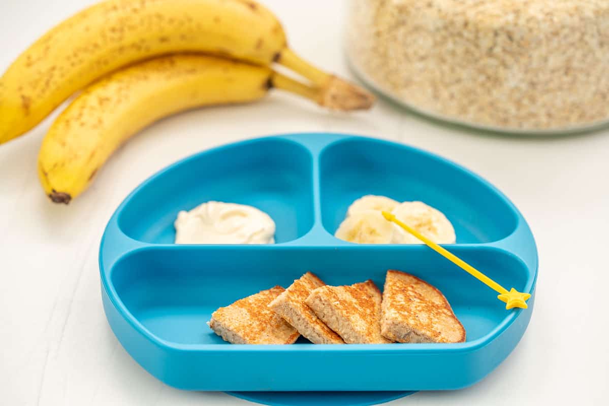 Banana oat pancakes sliced into triangle wedges, on a blue baby plate with banana slices and yoghurt.