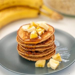 A stack of 5 pancakes on a blue plate with sliced banana and maple syrup.
