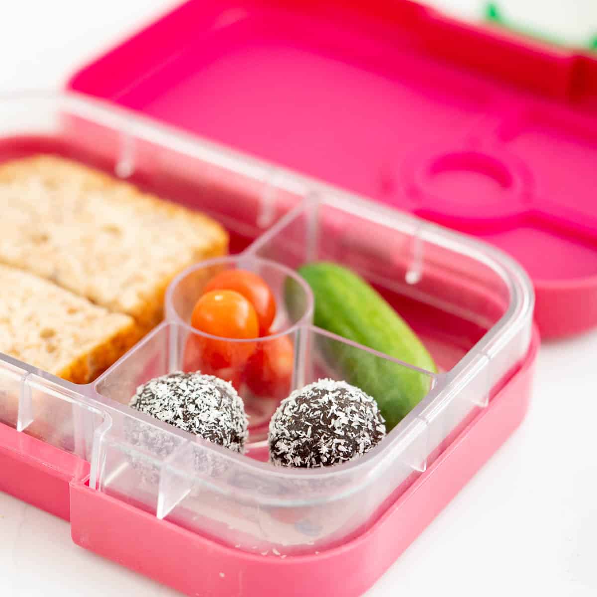 A pink bento box packed with chocolate bliss balls, cucumber, tomatoes and a sandwich.