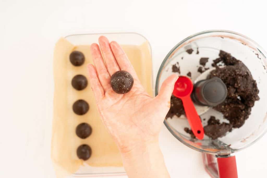 A women's hand holding a chocolate date ball above a container of date balls.