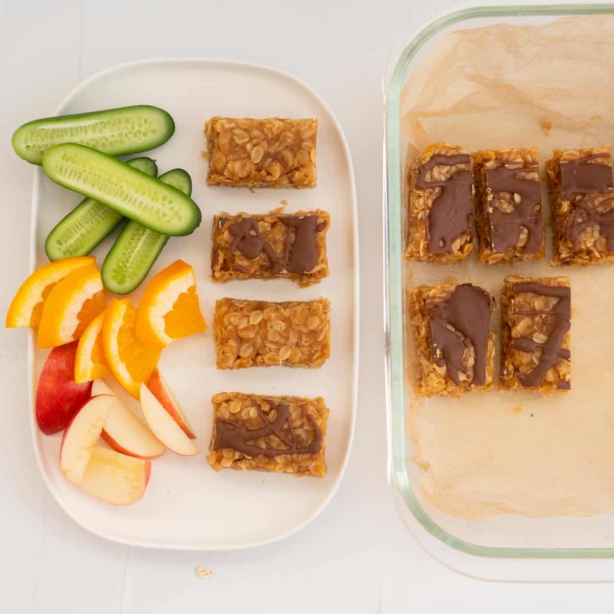 Peanut butter oat bars and fresh fruit and veggies on a white platter next to a container of bars ready to be refrigerated.