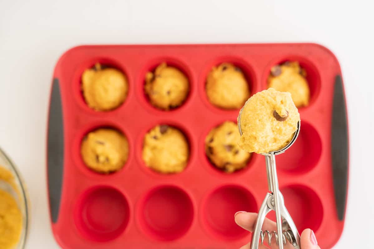 Woman's hand holding a scoop of muffin batter above a red silicone muffin tray.