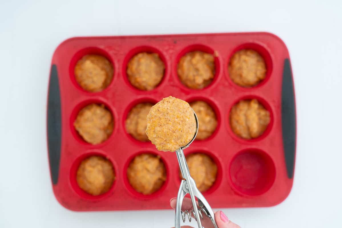 A scoop of sweet potato muffin batter being spooned into a red silicone muffin tray.