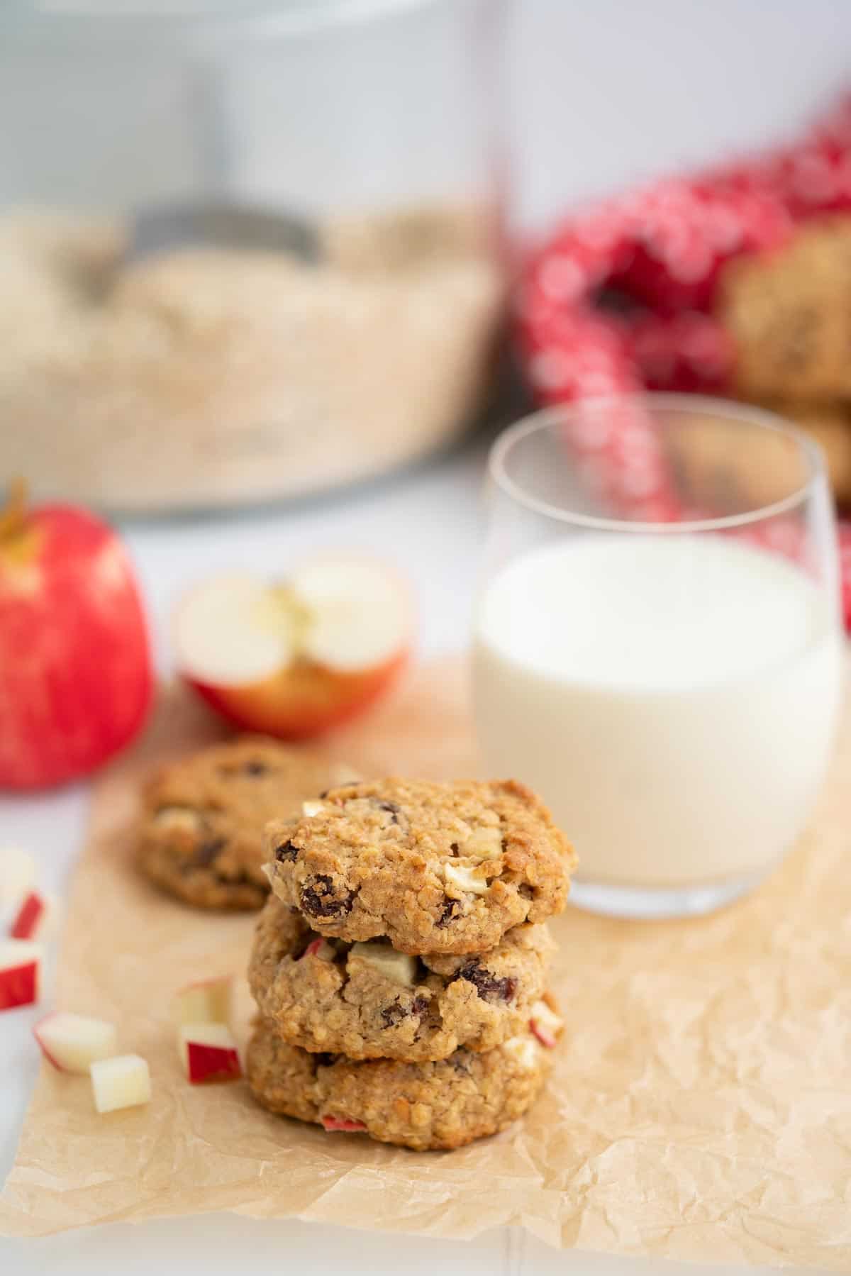 A stack of three oat meal cookies sitting on crinkled parchment paper with a glass of milk, apples and a jar of rolled oats in the background.