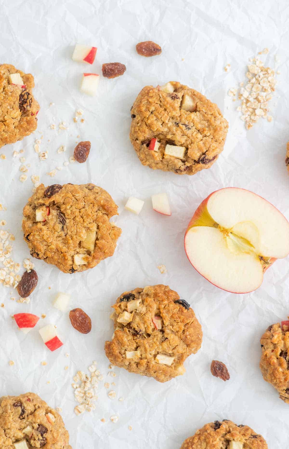 Oat cookies on white crinkled baking paper scattered with apple pieces, oats and raisins.
