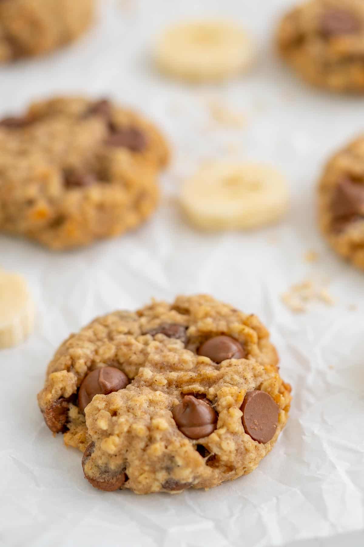 A banana oatmeal cookie on crinkled baking paper, with slices of banana and a scattering of rolled oats and chocolate drops.