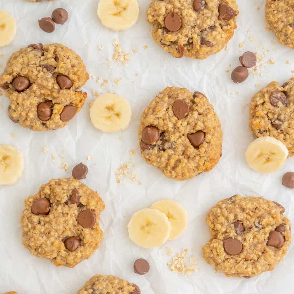 Banana oatmeal cookies laid out on baking paper, with slices of banana and a scattering of rolled oats and chocolate drops.