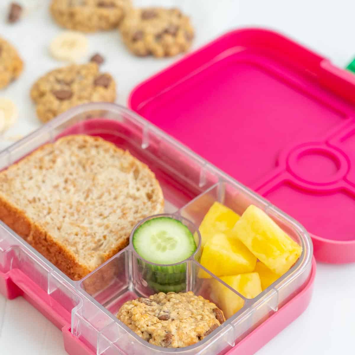 A pink bento style lunchbox, packed with a sandwich, pineapple, oatmeal cookie and cucumber slices.