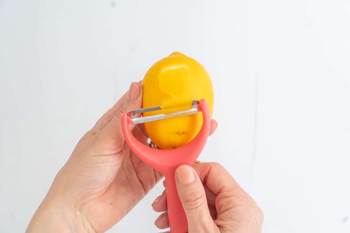 A large strip of lemon zest being removed from lemon with a pink potato peeler.