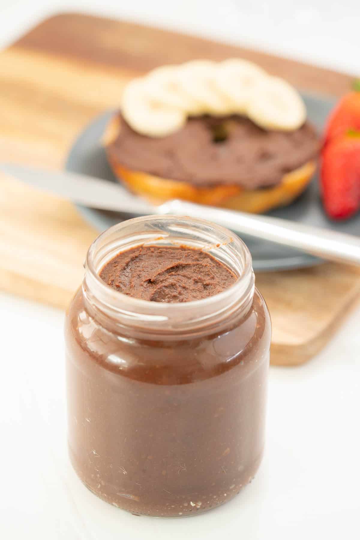 A jar of chocolate spread sitting in front of a bagel, plate and knife.