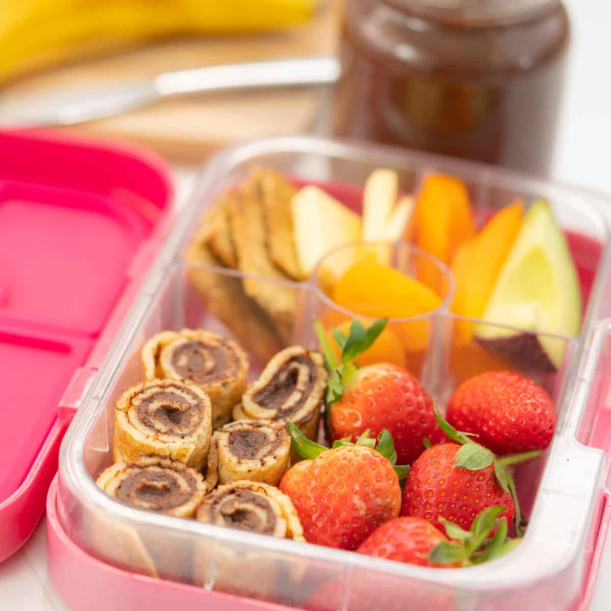 A pink bento box packed with brownie hummus pancakes, strawberries, crackers and cheese and avocado.