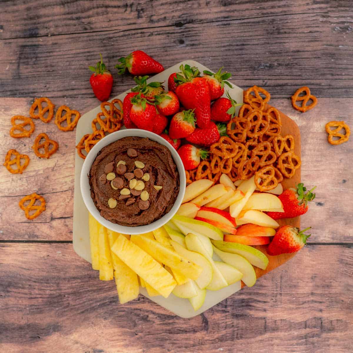 A platter of fresh fruit and pretzels with a bowl of chocolate hummus.