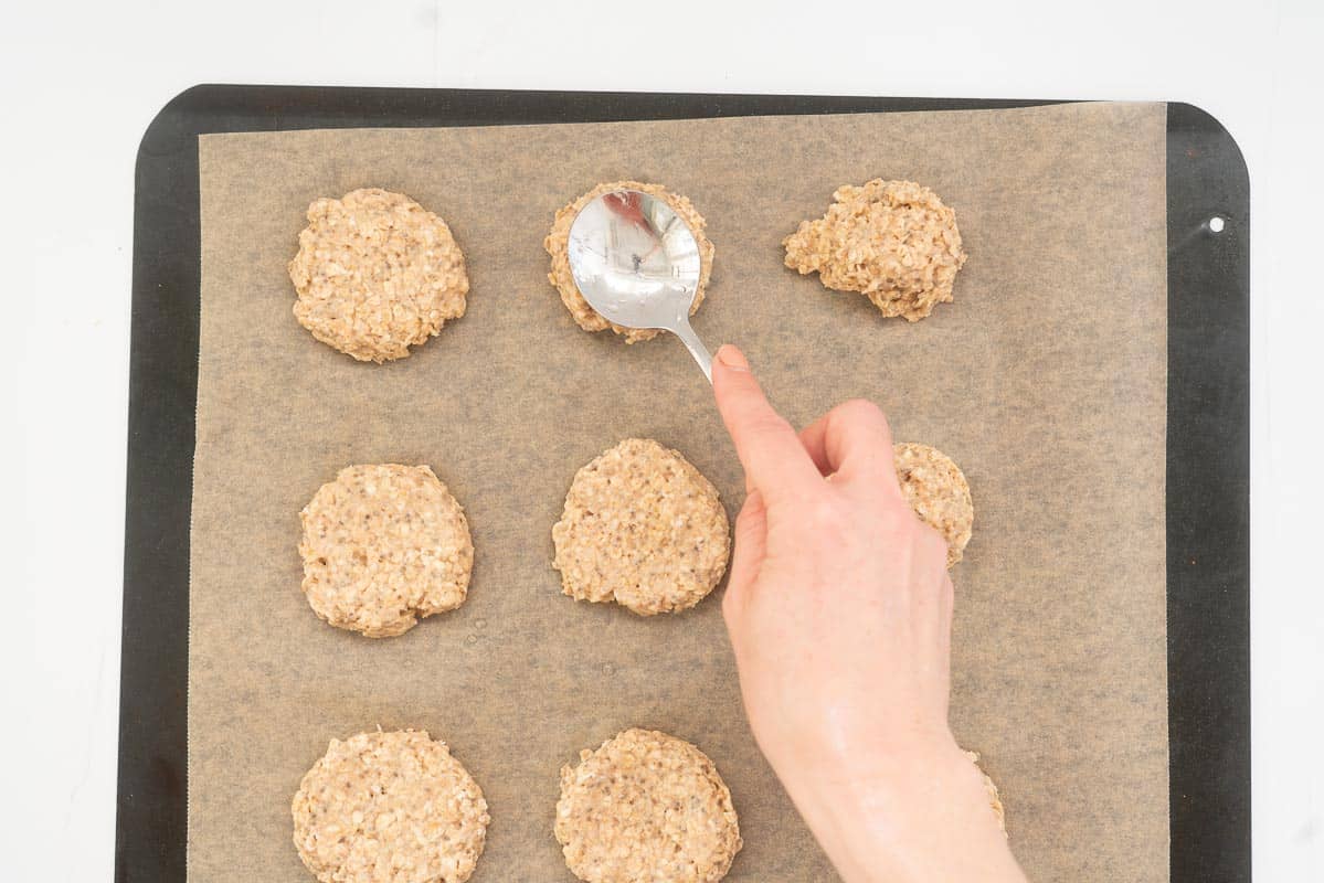 Balls of cookie dough on a baking paper lined tray being flattened with the back of a spoon.