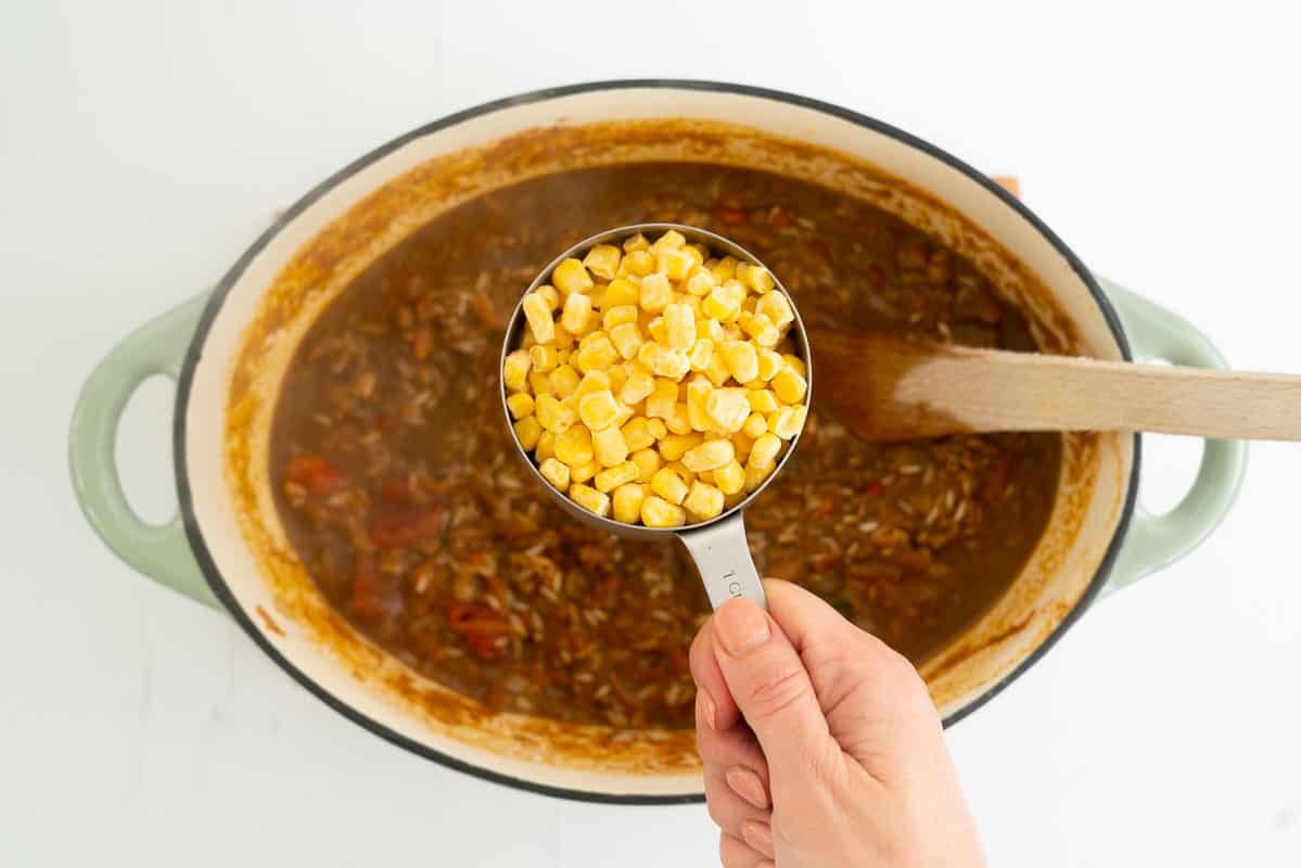 A cup full of frozen corn kernels held above a dutch oven of Mexican mince and rice.
