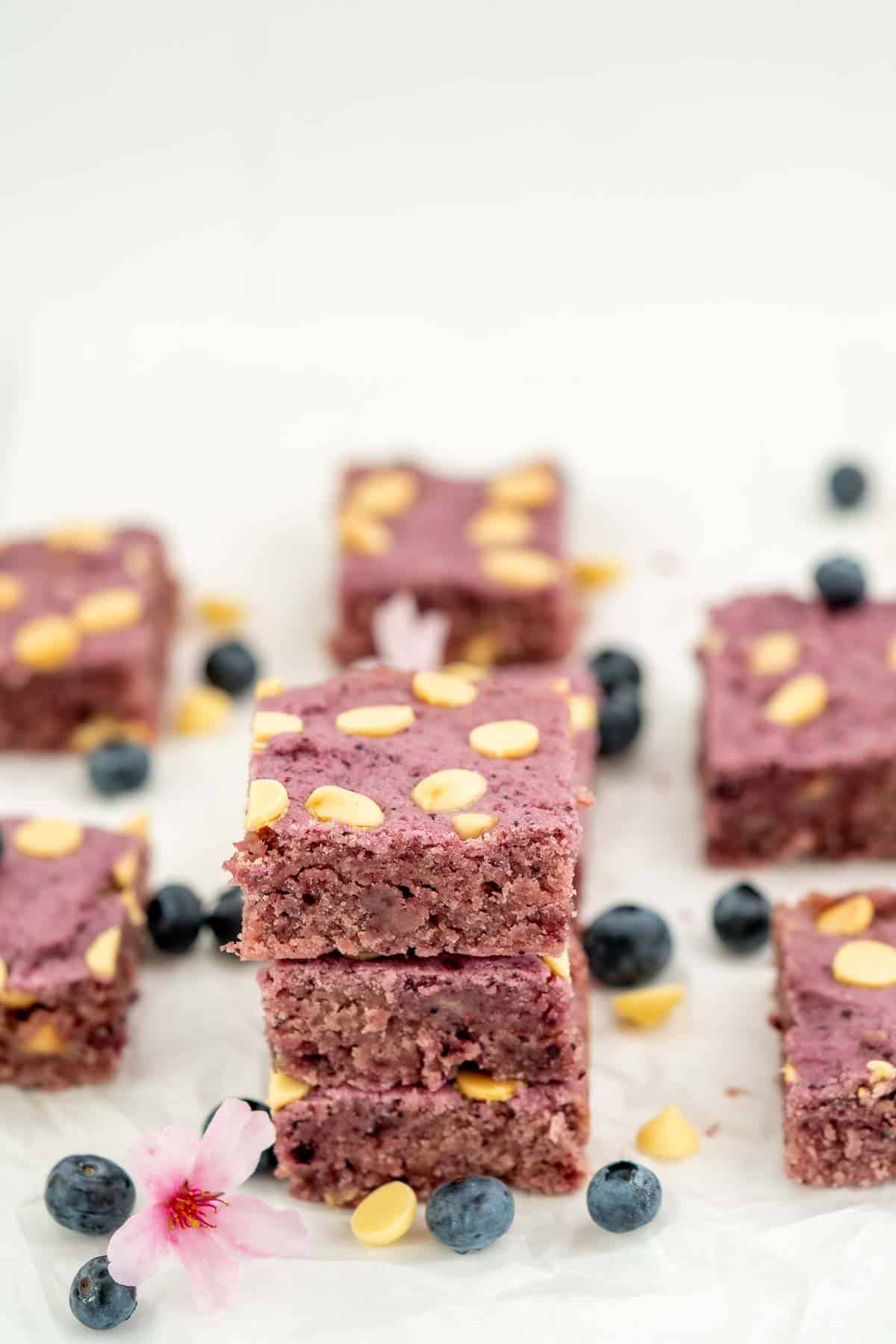 A stack of three blueberry cookie bars sitting on a white surface scattered with blueberries, flowers and white chocolate drops.