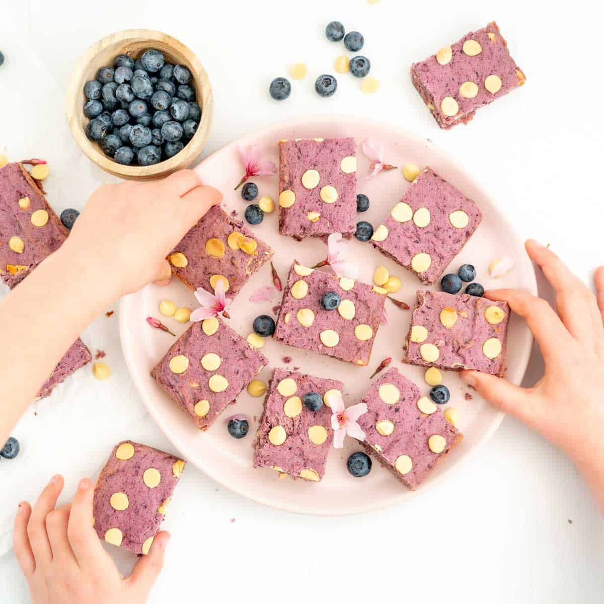 Three children's hands reaching for purple cookie bars sitting on a pink plate.
