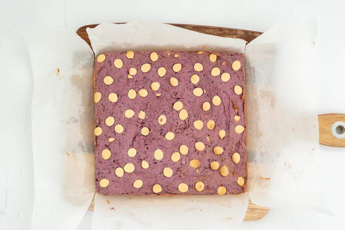 A square of baked blueberry cookie bars on a wooden chopping board ready to be sliced.
